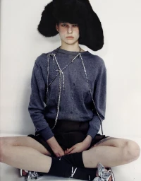 Model seated wearing a grey cardigan, oversized black fuzzy hat, rhinestone body jewelry and metallic shoes