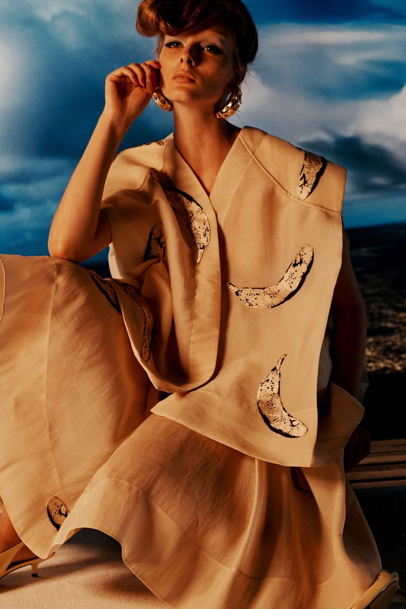 A woman in pale clothing with banana prints poses against a blurred outdoor backdrop, creating a fashion-forward and slightly surreal image.