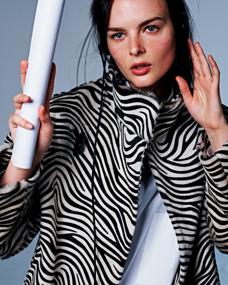 Adult female wearing zebra-striped coat holds rolled white paper. Close-up studio shot. Fashion editorial.
