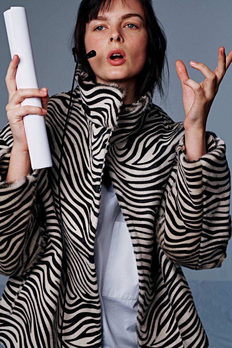Adult woman wearing a zebra print coat holds a white paper roll and gestures with her hand in a studio setting with grey background.