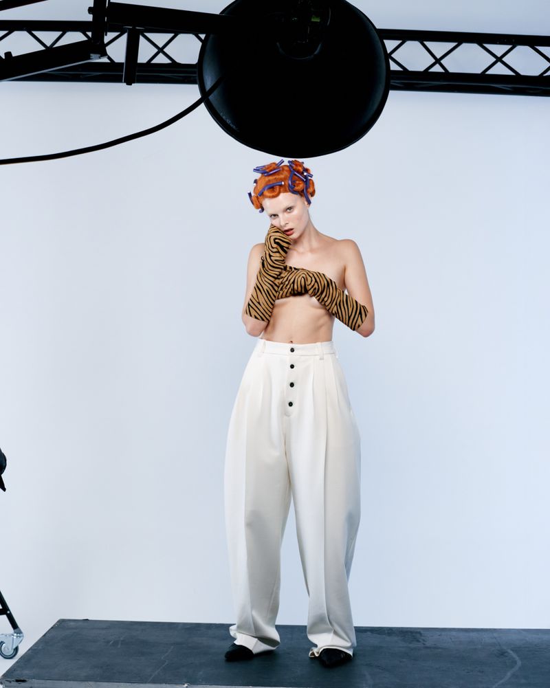 Fashion image of a topless woman in wide-legged pants and patterned gloves standing in a studio with hair rollers.