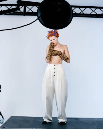 Fashion image of a topless woman in wide-legged pants and patterned gloves standing in a studio with hair rollers.
