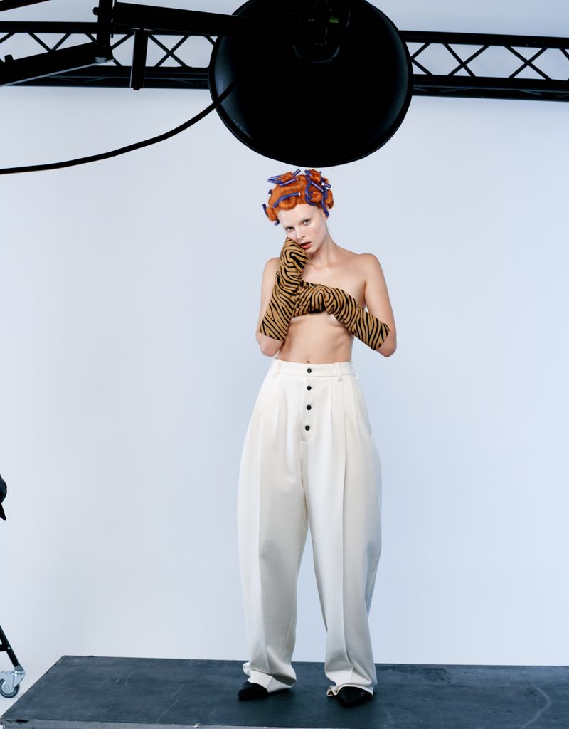 Nude model in oversized white pants and tiger-striped gloves poses in a photography studio with hair rollers and studio lighting.