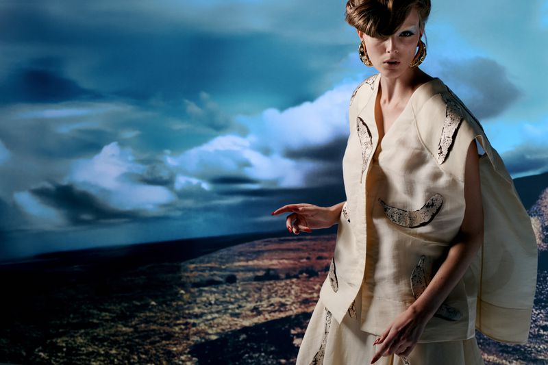 Adult woman in sleeveless top and long skirt with an outdoor landscape background featuring a cloudy sky and arid land.