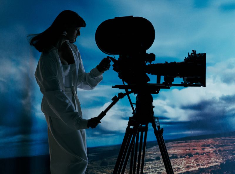 Silhouette of a woman in a white suit operating a vintage film camera on a tripod against a cloudy blue sky background.