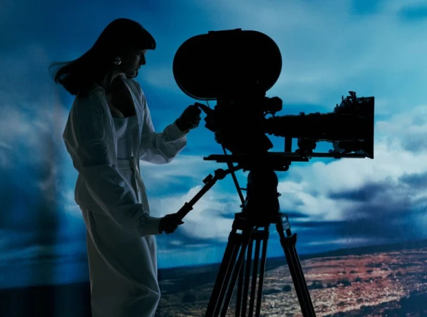 Silhouette of a woman in a white suit operating a vintage film camera on a tripod against a cloudy blue sky background.