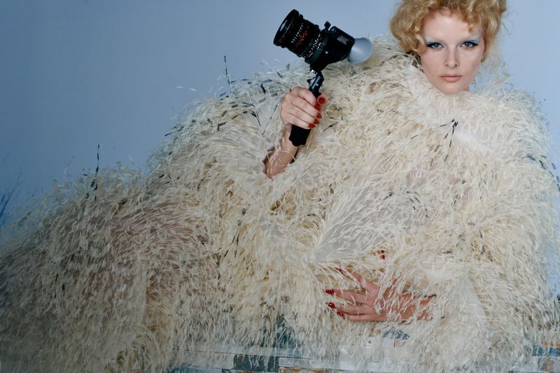 Fashion portrait of a woman with curly hair and a feather boa holding a black camera against a blue background. Medium shot, indoors.