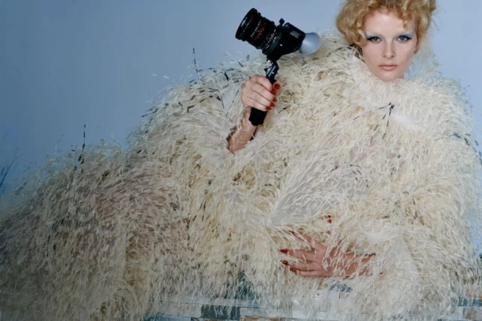 Fashion portrait of a woman with curly hair and a feather boa holding a black camera against a blue background. Medium shot, indoors.