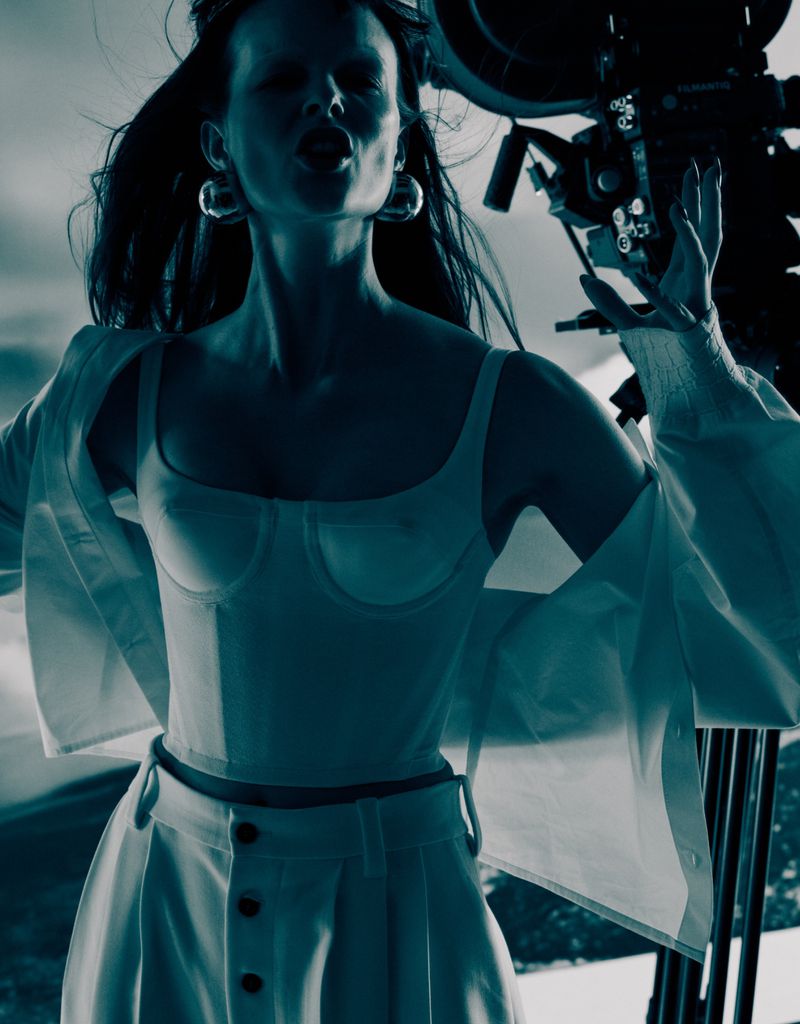Model Aylah Peterson wearing white top and trousers with large earrings in front of camera gear for Sixteen Journal