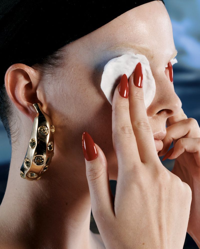 Adult female cleaning makeup off face using cotton pad, gold earrings, red nails, black head covering in indoor setting.