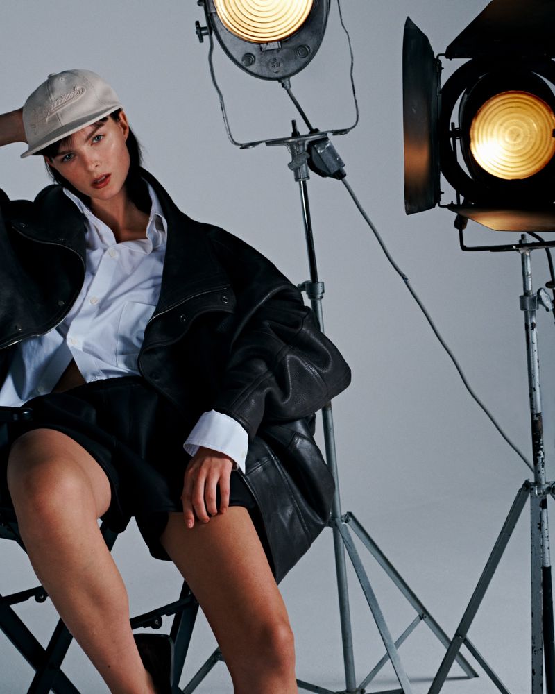 Brunette woman poses in a photo studio with black shorts, leather jacket, white shirt, and cap, sitting next to film production lights.