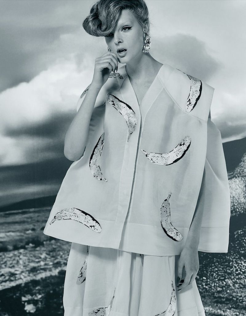 Elegant woman wearing white vest and skirt with banana pattern, silver statement earrings, fashionable updo against a cloudy background.