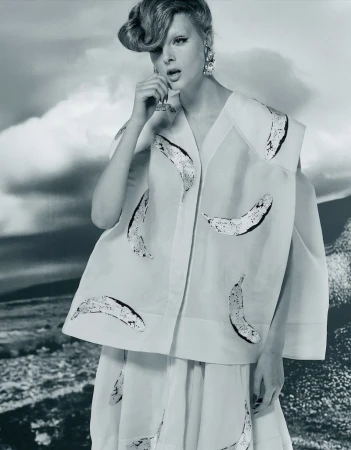 Elegant woman wearing white vest and skirt with banana pattern, silver statement earrings, fashionable updo against a cloudy background.