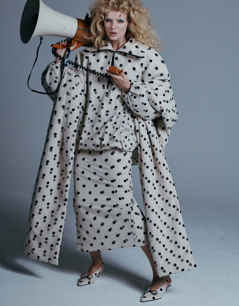 Fashionable woman in polka dot outfit poses with a retro bullhorn. Beige and black ensemble with voluminous sleeves in a gray studio.