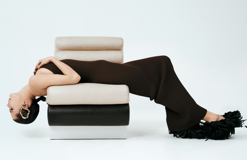 Adult female in brown dress reclines on modern chair against a white backdrop, wearing gold earrings and feathered shoes in a studio setting.