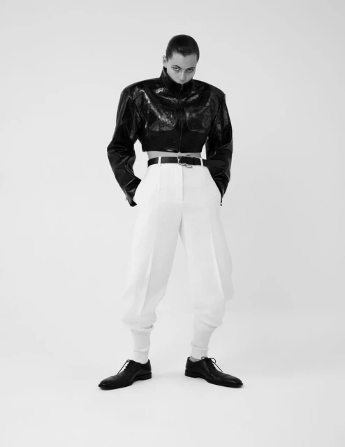 Full-length portrait of a person in a short, black patent leather jacket and white balloon pants against a white studio background.