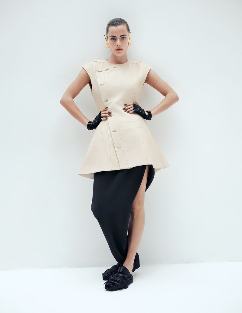 Full-length shot of a woman in a beige dress, black skirt, black gloves, and black platform shoes, posing against a white backdrop.