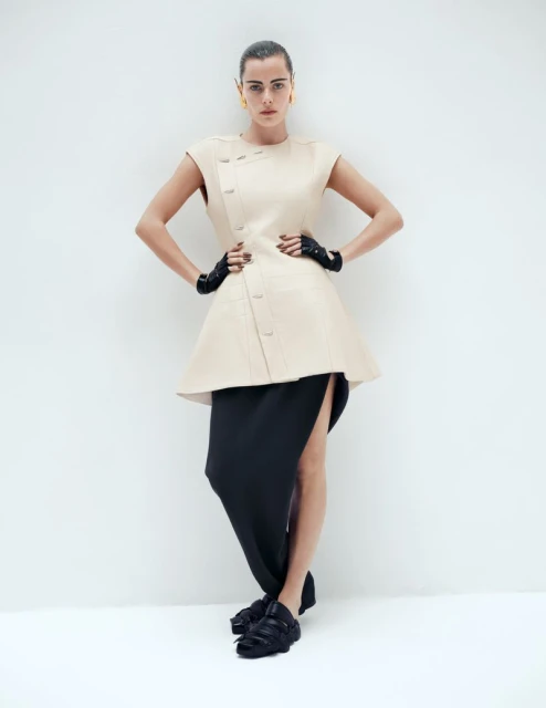 Full-length shot of a woman in a beige dress, black skirt, black gloves, and black platform shoes, posing against a white backdrop.