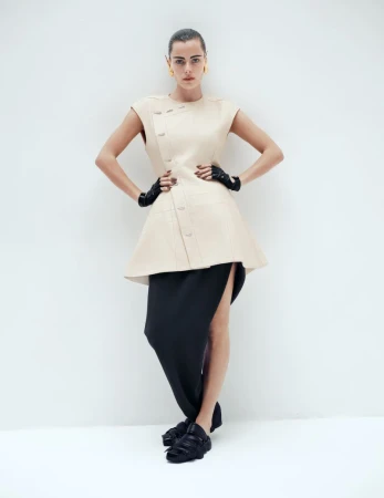 Full-length shot of a woman in a beige dress, black skirt, black gloves, and black platform shoes, posing against a white backdrop.