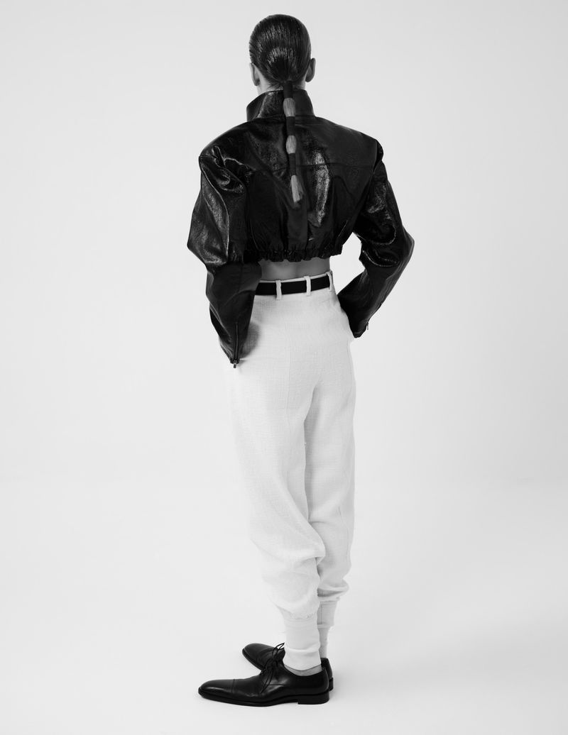 Back view of a slender adult model in a black leather crop top, white pants, black belt and shoes, on a solid white background.