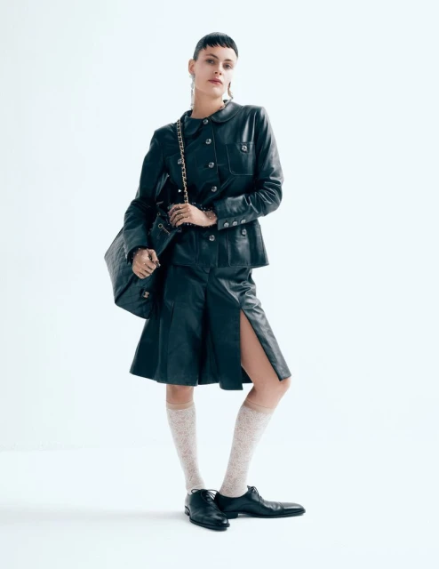 Fashion editorial shot of a woman in a black leather suit and sheer socks, accessorized with a chain strap bag and earrings, on a white background.