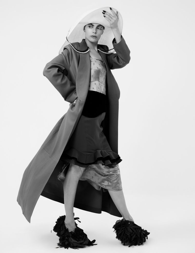 Fashion model in a long coat, hat, and unique shoes posing in a studio with a neutral background, showcasing modern editorial style.