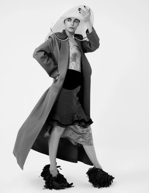 Fashion model in a long coat, hat, and unique shoes posing in a studio with a neutral background, showcasing modern editorial style.