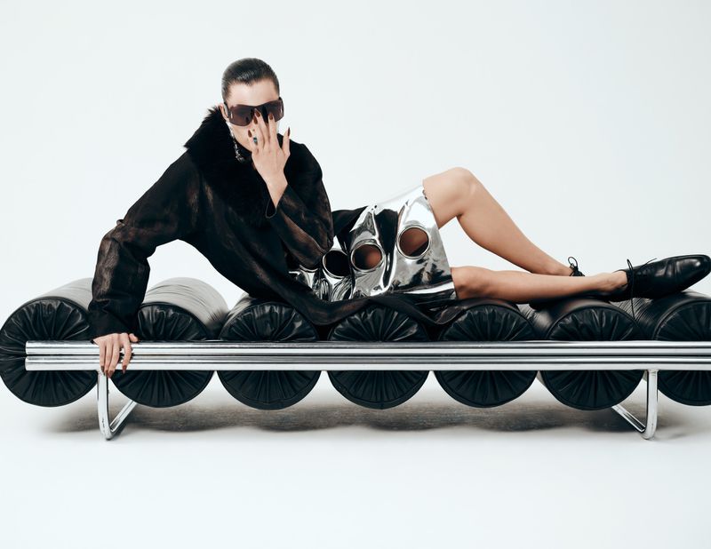 A woman in a black coat and shiny silver skirt reclines on a black cylindrical couch with chrome legs in a white studio setting.