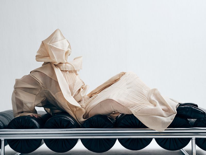 Fashion photography showcasing a person reclining on a bench, wearing a textured, ivory-colored couture outfit against a white backdrop.