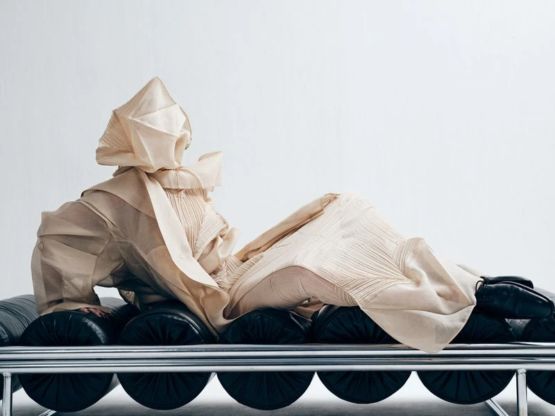 Fashion photography showcasing a person reclining on a bench, wearing a textured, ivory-colored couture outfit against a white backdrop.