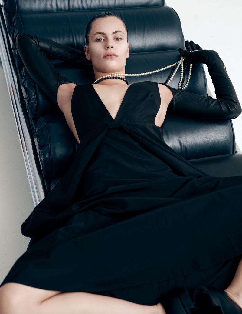 A woman in a black dress and long gloves sits on a black leather chair, holding a pearl necklace against a white background.
