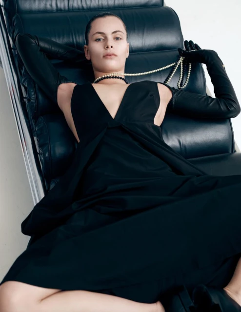 A woman in a black dress and long gloves sits on a black leather chair, holding a pearl necklace against a white background.