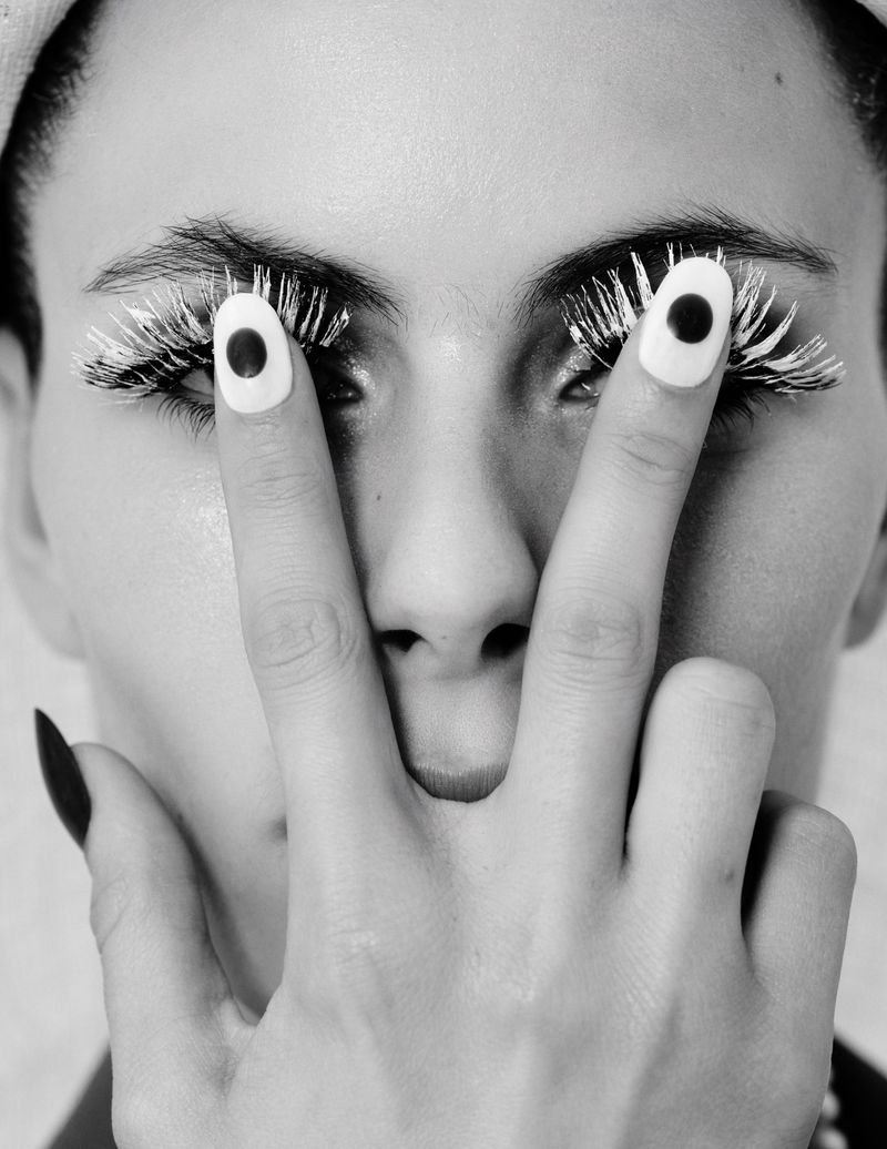 Close-up black and white portrait of a woman with white painted eyelashes and polka dot nail art holding her hand up to her face.