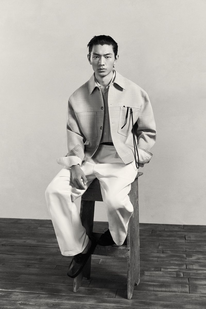 Monochrome studio shot of a male model in a collared shirt and patterned jacket, seated on a wooden stool against a plain backdrop.