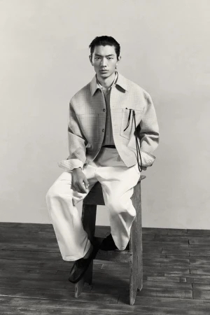 Monochrome studio shot of a male model in a collared shirt and patterned jacket, seated on a wooden stool against a plain backdrop.