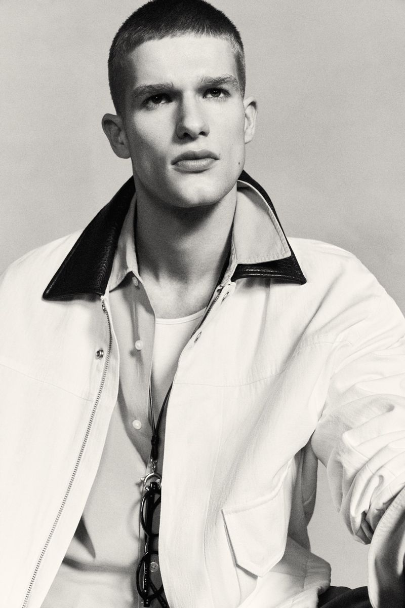 Portrait of a male model wearing a light-colored jacket with dark collar and a button-down shirt against a neutral studio backdrop.