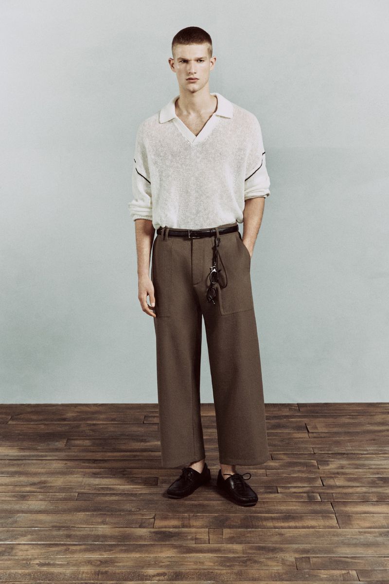 Full-length fashion portrait of a young adult male model wearing a polo shirt and wide-leg trousers, standing in a studio setting.