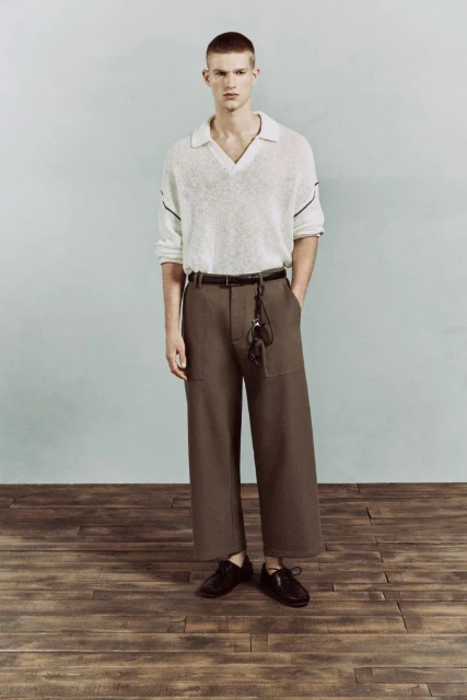 Full-length fashion portrait of a young adult male model wearing a polo shirt and wide-leg trousers, standing in a studio setting.