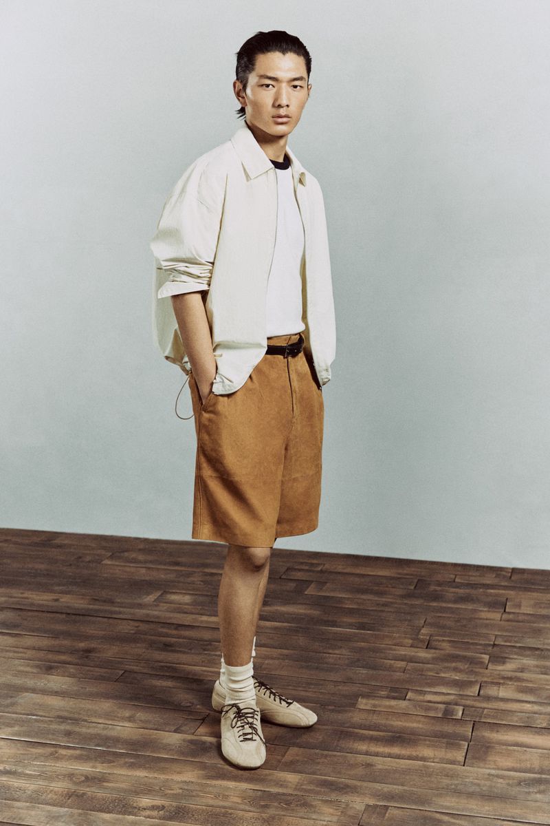 Full shot of a stylish Asian male model in a beige jacket and brown shorts, posing in a studio with a wood floor.