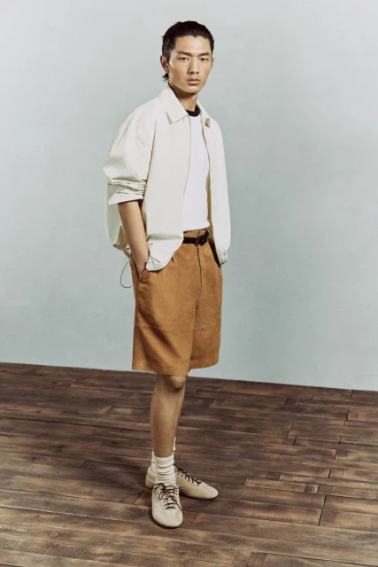 Full shot of a stylish Asian male model in a beige jacket and brown shorts, posing in a studio with a wood floor.
