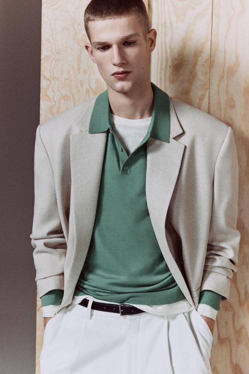 Young man in a beige blazer and white pants posing against a plywood wall, fashion photography, studio lighting, modern style.