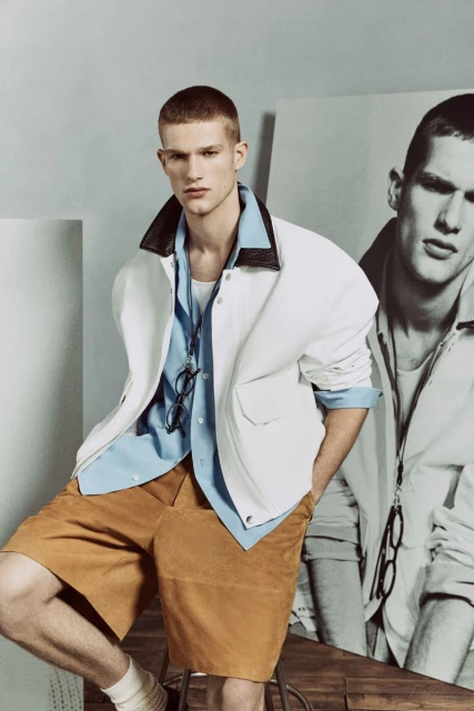 A fair-skinned adult male model in a white jacket and tan shorts poses indoors against a background with an image of another man.