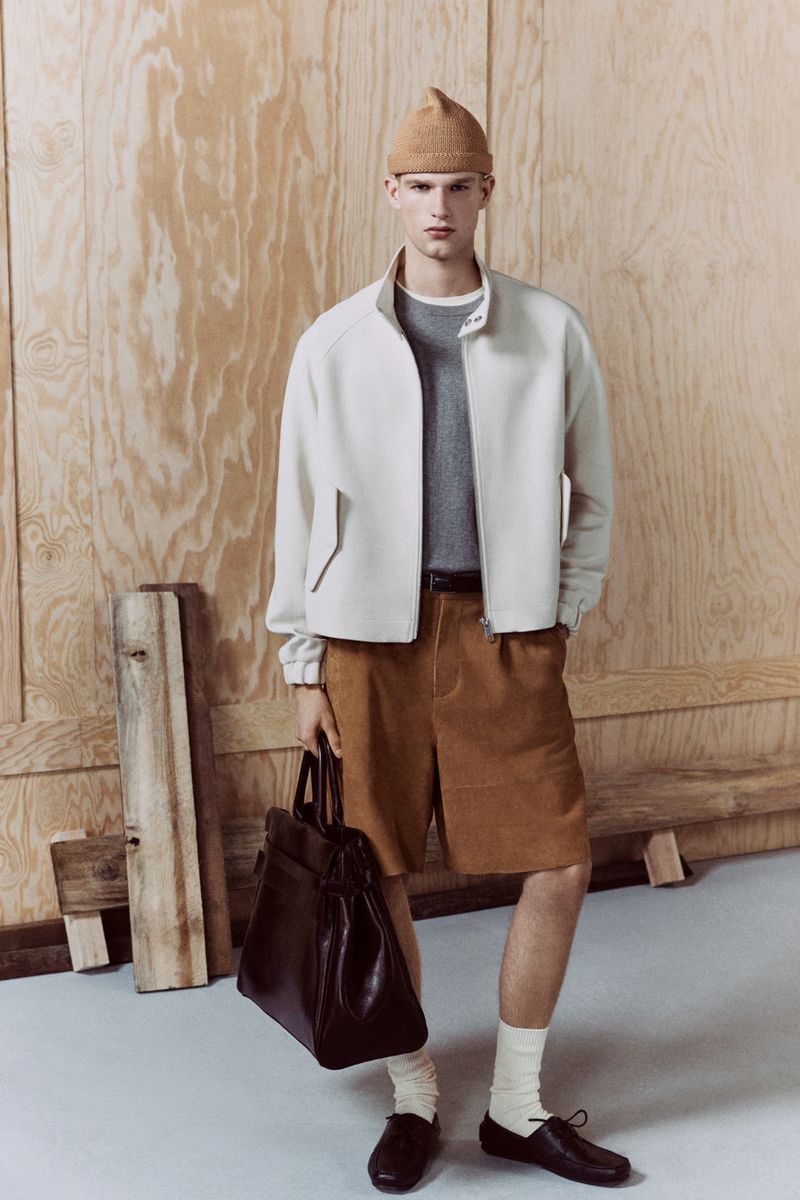 Stylish man in beige jacket and brown shorts holding a dark bag stands against a plywood background in a fashion studio setting.