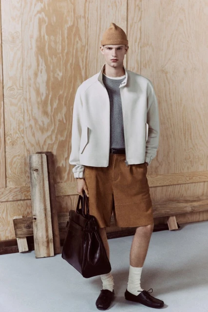 Stylish man in beige jacket and brown shorts holding a dark bag stands against a plywood background in a fashion studio setting.
