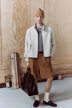 Stylish man in beige jacket and brown shorts holding a dark bag stands against a plywood background in a fashion studio setting.