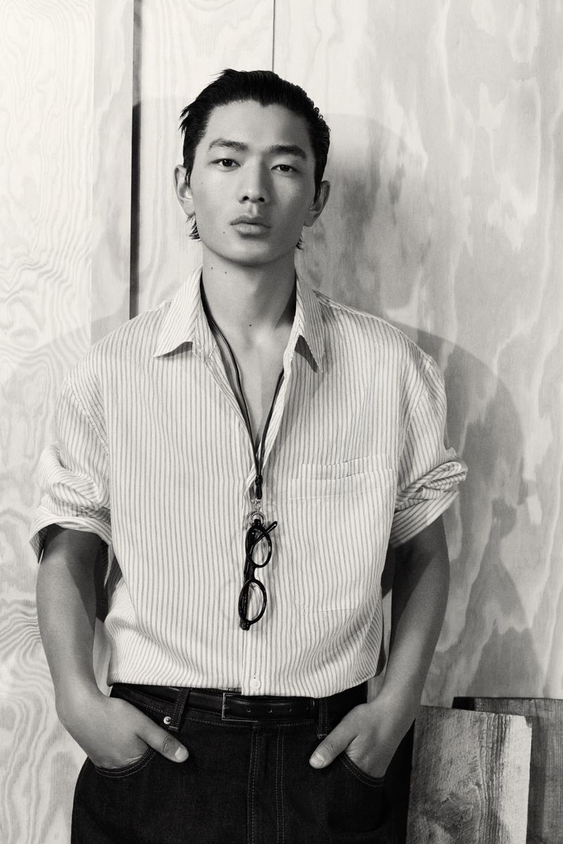 Monochrome portrait of a young Asian man wearing a striped shirt and jeans, standing against a wood panel background with glasses.