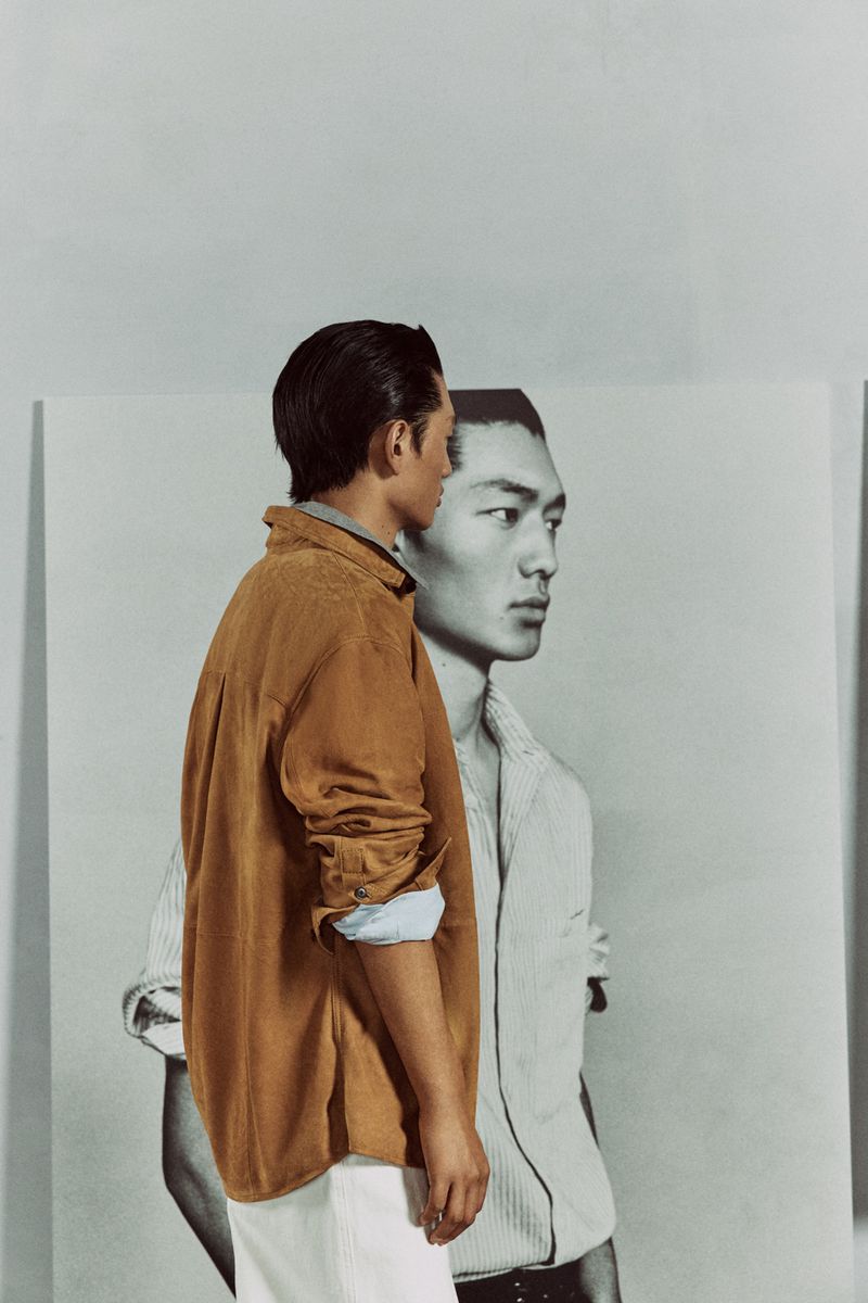 Man in an open-front brown shirt stands near a large black-and-white portrait of another man in a minimalist off-white studio.