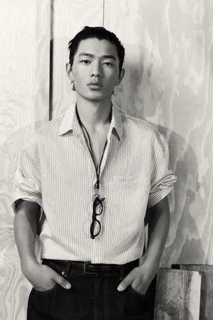 Monochrome portrait of a male model wearing a striped button-down shirt and dark pants against a wooden panel wall