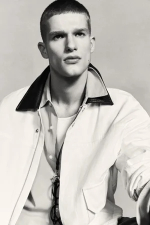 Portrait of a male model wearing a light-colored jacket with dark collar and a button-down shirt against a neutral studio backdrop.