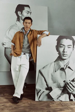 A man wearing a tan overshirt, grey polo, and white trousers poses next to large portrait prints for Zara Man Spring Neutrals.